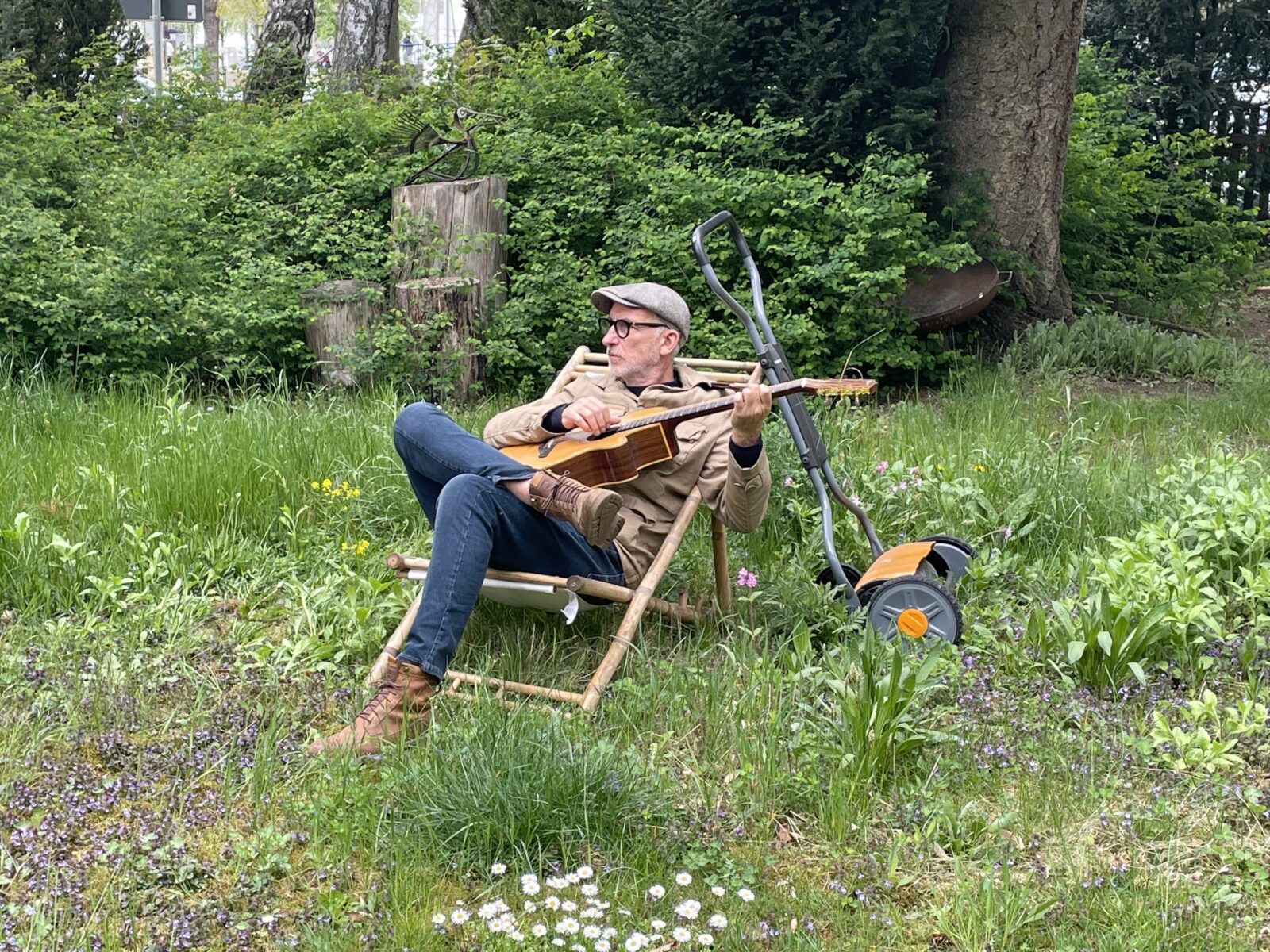 Umweltreferent Michael Zott sitzt mitten in einer ungemähten Wiese mit Gitarre im Liegestuhl. Am Liegestuhl lehnt ungenutzt ein Rasenmäher.